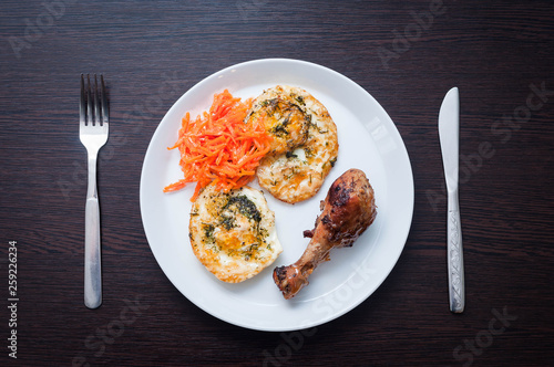 The perfect morning meal for a healthy lifestyle of two fried eggs with greens, chicken leg and Korean carrot. On a white plate, on a black table with a fork and knife
