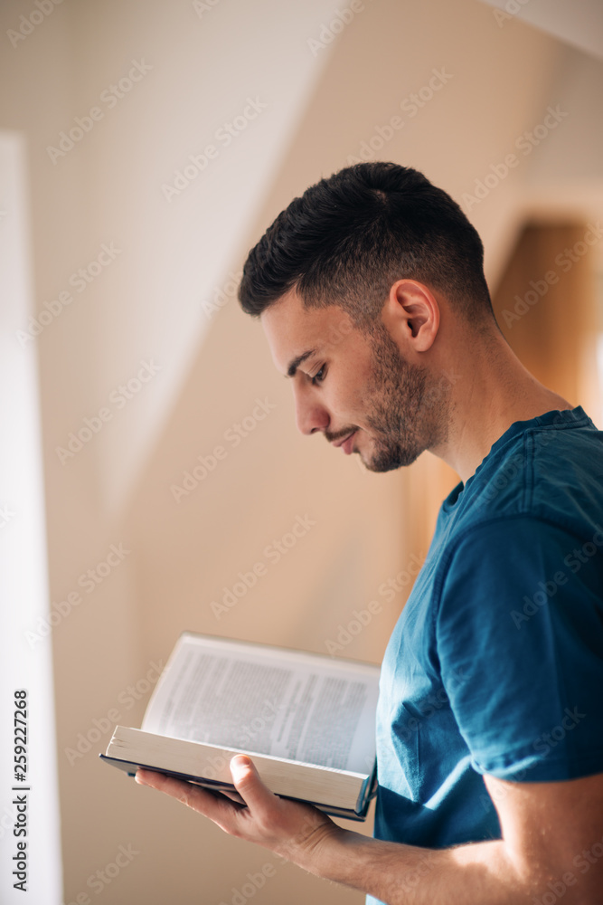 Student reading a book while standing and walking in the room next to the windows. Student reading a book while standing and walking in the room next to the windows.