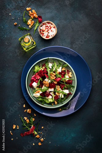 Beet or beetroot salad with fresh arugula, radicchio, soft cheese and walnuts on plate with fork, dressing and spices on blue kitchen table background, copy space, top view