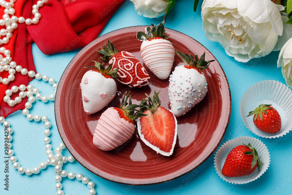 Fototapeta premium Ripe strawberries in white chocolate glaze lay on a plate close to white flowers and pearl beads on a turquoise background