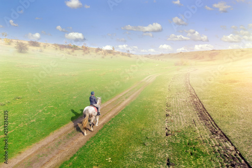 Young female equestrian riding horse along rural countryside. Rider on horseback going through green hillside.Travelling along. Aerial back view