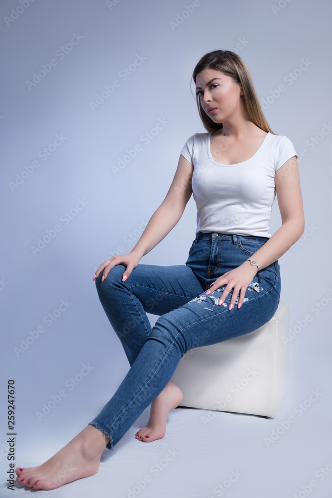 Cute young woman in blue jeans and bare feet sitting on white cube stool in studio and isolated on white blue background. Female fashion portrait concept. Close up