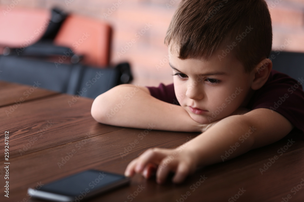 Sad little boy with mobile phone sitting at table indoors Sad little boy with mobile phone sitting at table indoors