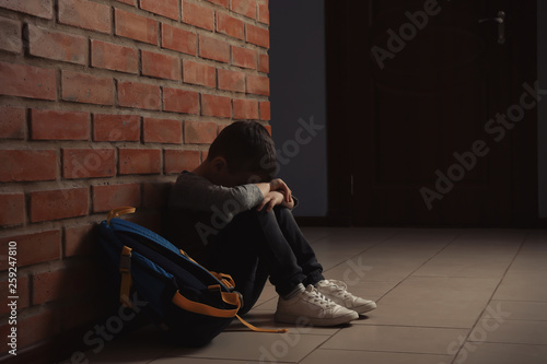 Photography Sad little boy with backpack sitting on floor near brick wall indoors