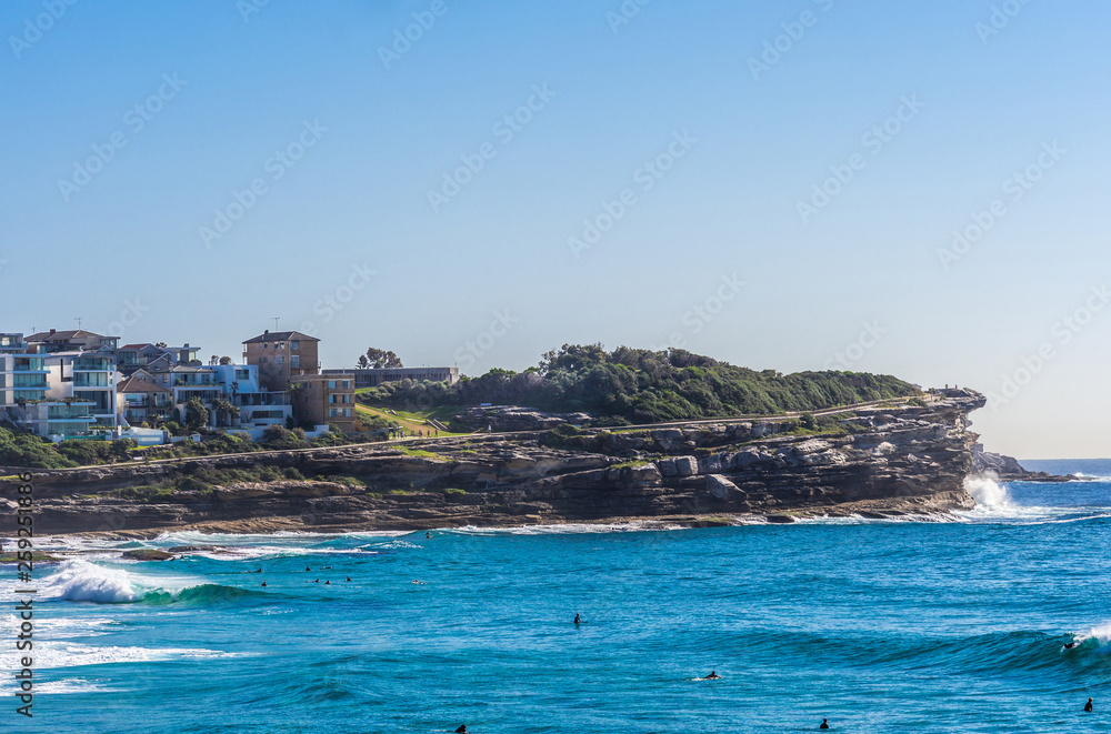Sydney, Australia - February 11, 2019: Wider shot of Gray with green vegetation on top North Cliff point of Bronte Beach, blue sea water with swimmers, under light blue sky, seen from South cliff. 