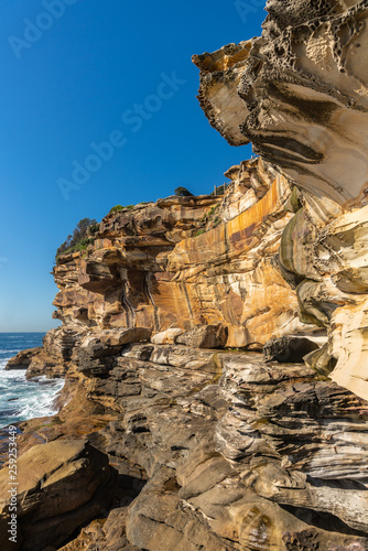 Sydney, Australia - February 11, 2019: Spectacular rock outcrop at Bronte Beach South cliffs, made by erosion.. Yellows and browns under blue sky. Sponge-like borders of shell like plates sticking out