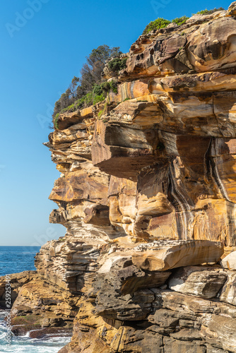 Sydney, Australia - February 11, 2019: Closeup of Spectacular rock outcrop at Bronte Beach South cliffs, made by erosion.. Yellows and browns under blue sky. Sponge-like borders of shell like plates s