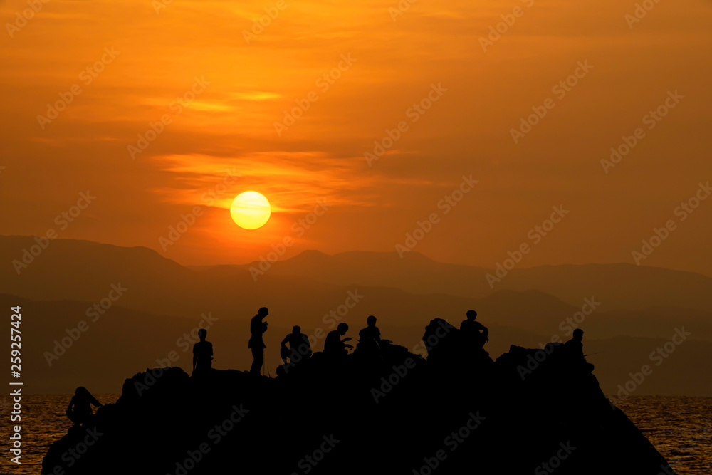 Silhouette of tourists Fishing on the rocks in sea and Golden light of sunrise behind the mountains.