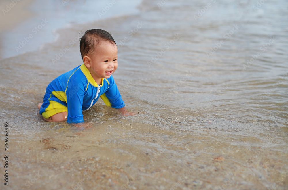 Happy baby boy in swimming suit having fun playing sea waves and water on the beach.