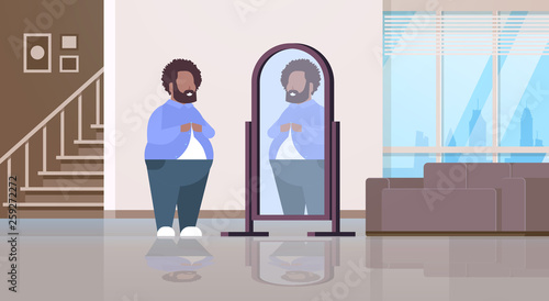 sad overweight man looking at himself reflection in mirror african american over size guy buttoning shirt unhealthy lifestyle obesity concept modern apartment interior flat full length horizontal