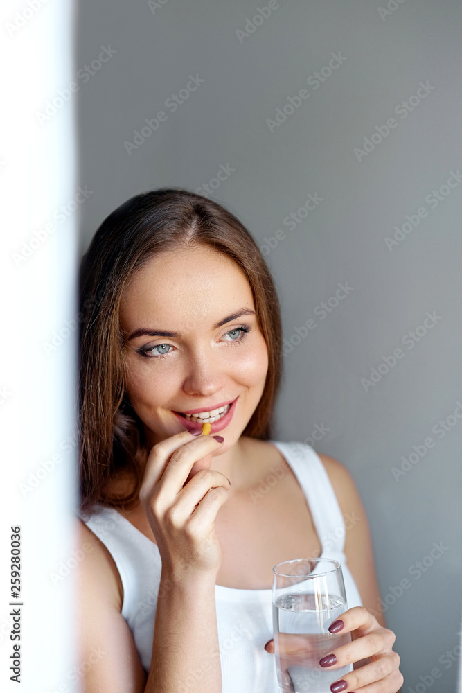 Young beautiful girl or woman take a pill or tablet with a glass of water near the window in white t-shirt Young beautiful girl or woman take a pill or tablet with a glass of water near the window in white t-shirt