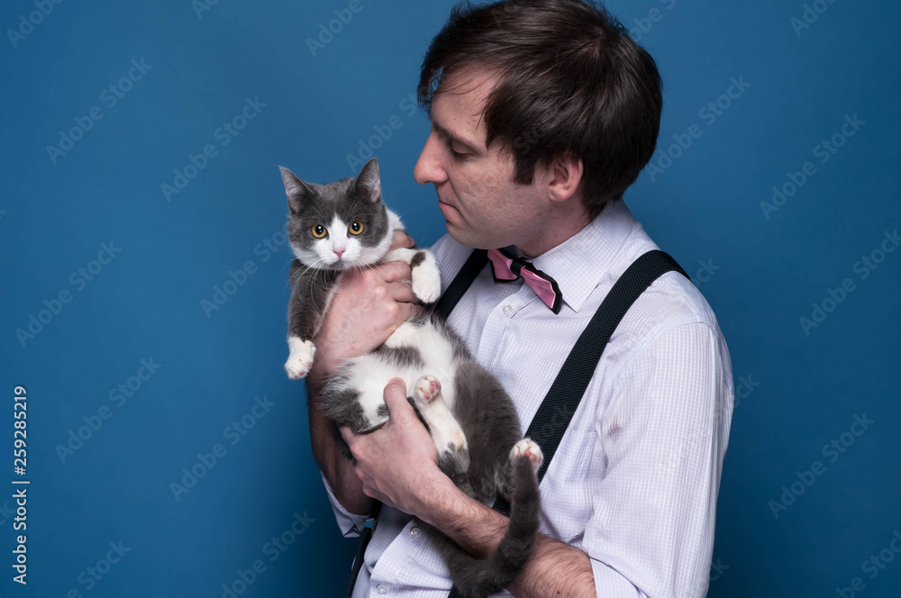 handsome man in shirt, suspender and pink bow tie holding and looking to cute grey cat with white paws on blue background with copy space handsome man in shirt, suspender and pink bow tie holding and looking to cute grey cat with white paws on blue background with copy space