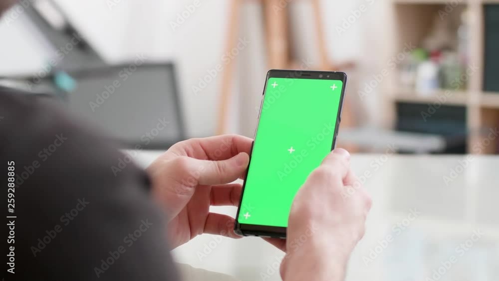 Man sits at a desk holds his smartphone with green screen on. Looking at the phone screen while at work. Office concept. Man sits at a desk holds his smartphone with green screen on. Looking at the phone screen while at work. Office concept.