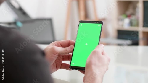 Man sits at a desk holds his smartphone with green screen on. Looking at the phone screen while at work. Office concept.