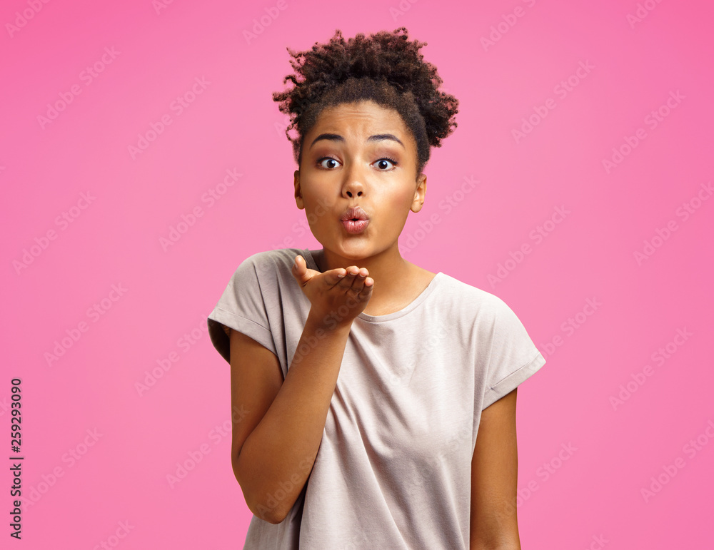 Pretty girl makes air kiss into camera, expresses her love. Photo of african american girl wears casual outfit on pink background. Emotions and pleasant feelings concept. Pretty girl makes air kiss into camera, expresses her love. Photo of african american girl wears casual outfit on pink background. Emotions and pleasant feelings concept.