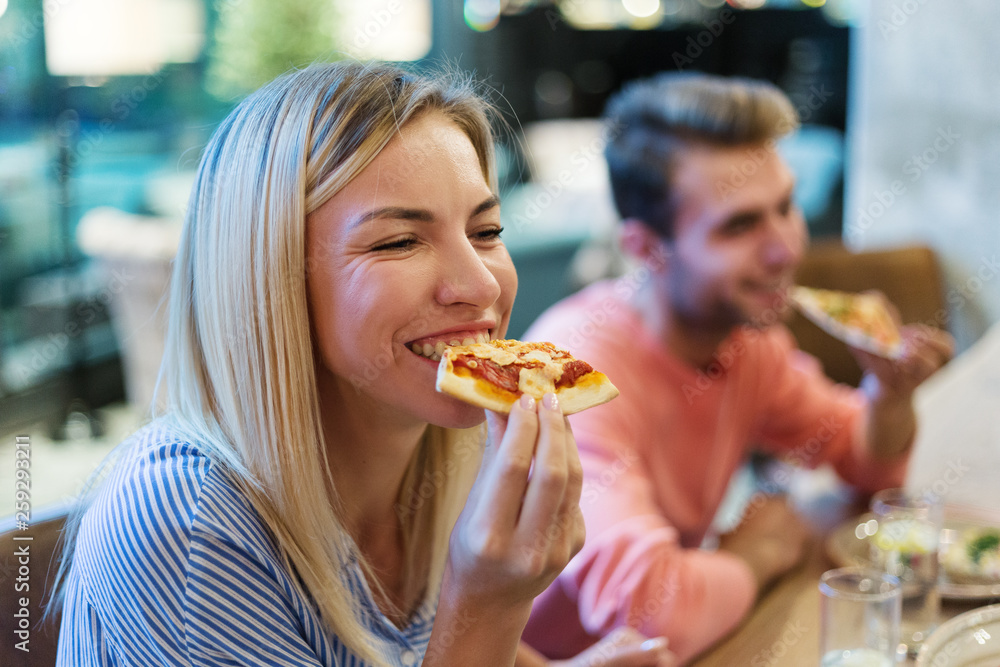 Joyful blonde woman enjoying eating delicious pizza at dinner in Italian restaurant, her male friend in background