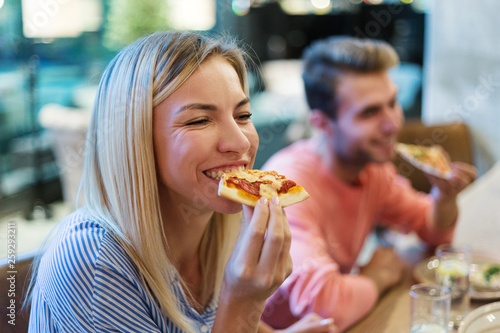Joyful blonde woman enjoying eating delicious pizza at dinner in Italian restaurant  her male friend in background