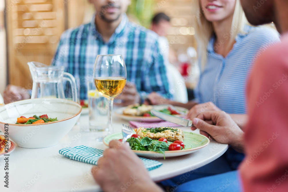 Close-up of fresh healthy dishes and glass of wine on dining table and defocused friends having dinner in restaurant