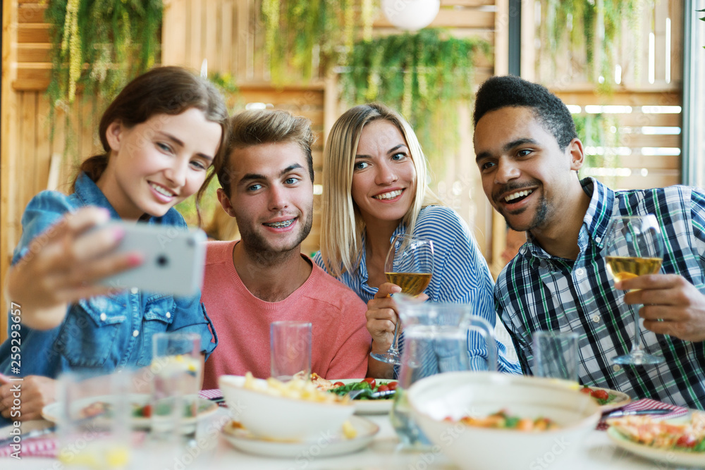 Group of four young people gathering for dinner in restaurant and posing together for selfie on smartphone Group of four young people gathering for dinner in restaurant and posing together for selfie on smartphone