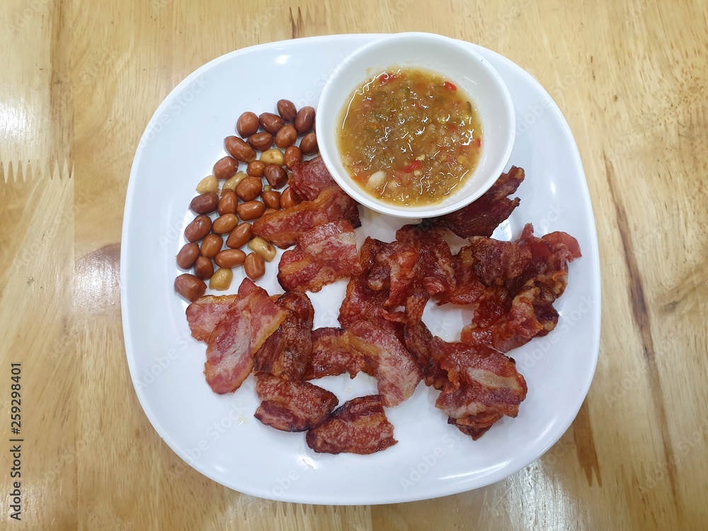 Top view of fried bacon and peanuts with spicy sauce in white plate on wooden table in restaurant