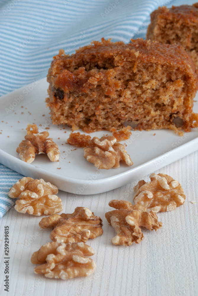Vertical close-up of carrot cake with nuts on white plate. On white wooden background and blue kitchen cloth with white stripes Vertical close-up of carrot cake with nuts on white plate. On white wooden background and blue kitchen cloth with white stripes
