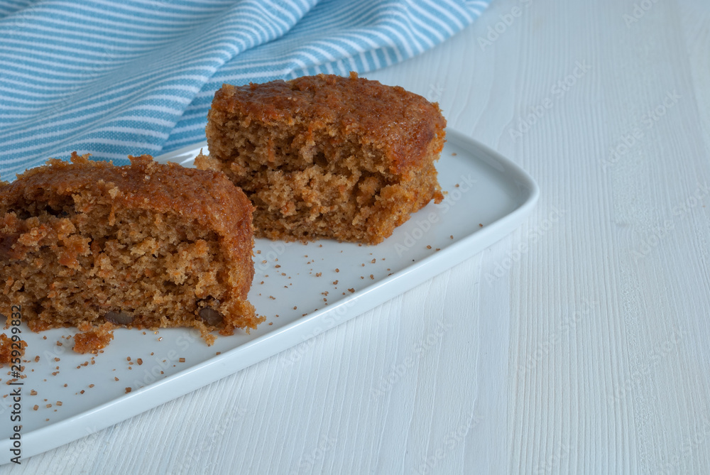 Horizontal close-up of carrot sponge cake with brown sugar. On white plate, white wood background and blue kitchen towel with white stripes with copy space Horizontal close-up of carrot sponge cake with brown sugar. On white plate, white wood background and blue kitchen towel with white stripes with copy space