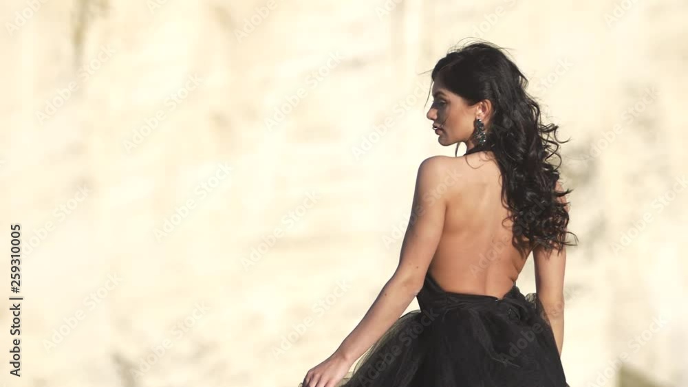 Back of young woman in black evening dress at backdrop of cliff Wind blow her black hair Back of young woman in black evening dress at backdrop of cliff Wind blow her black hair