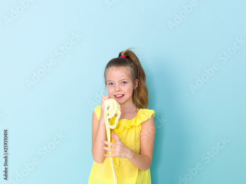 beautiful and cool and blond 9 year old girl is playing with yellow slime in front of blue background © epiximages