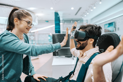 Young cheerful charming multicultural couple trying out virtual reality technology in tech store. Man wearing headset and sitting in chair while woman standing in front of him.