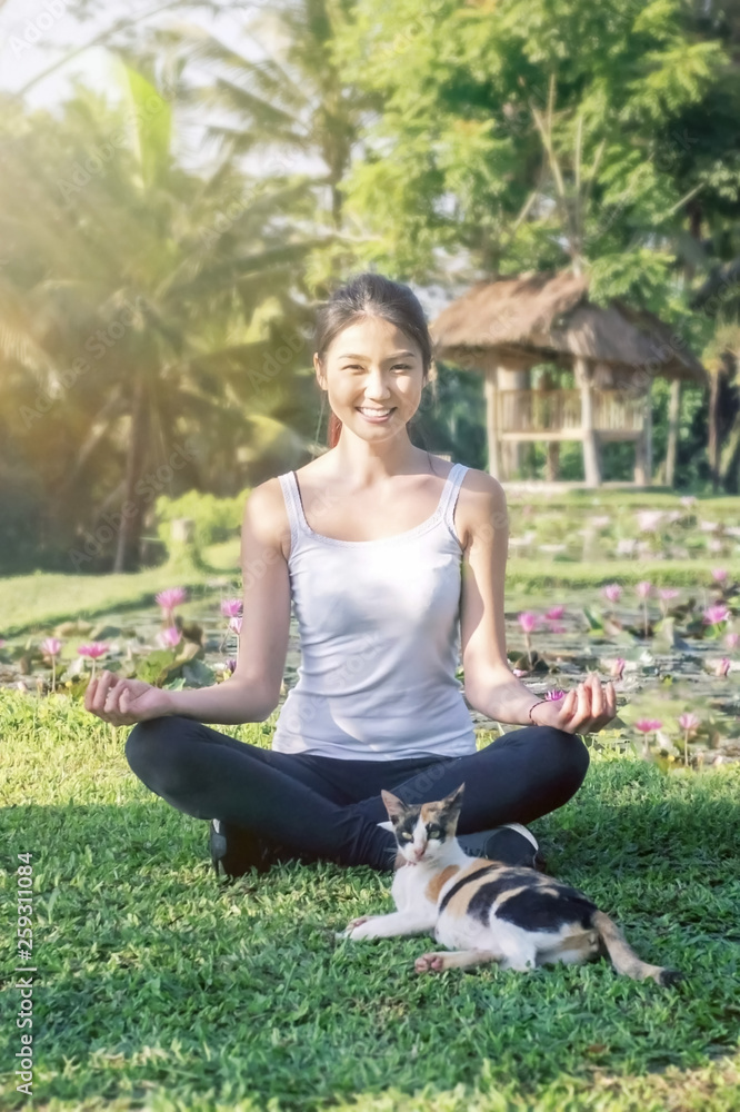Asian girl in the morning light sitting in the lotus position with a cat on the background of a lotus pond