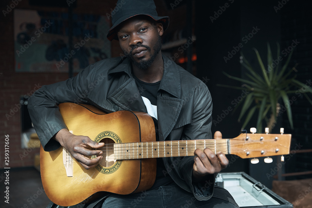 Obraz premium Musician in black trench coat and bucket hat sitting on bed and playing guitar. Songwriter in loft room. Fashion 2019