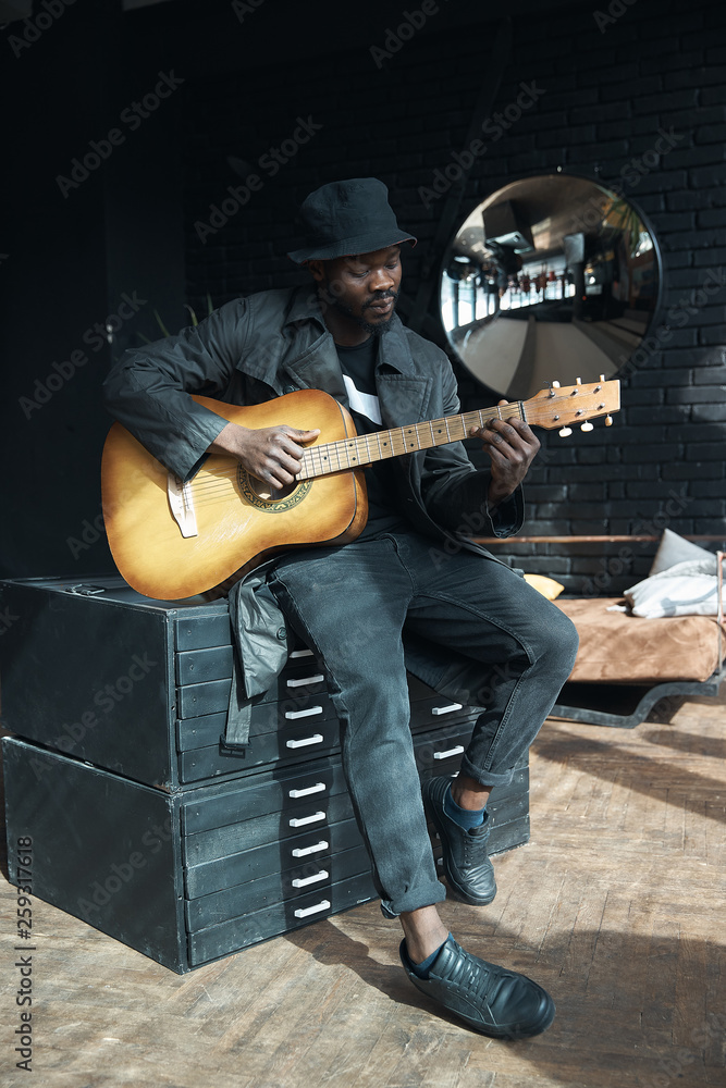 Musician in black trench coat and bucket hat sitting on bed and playing  guitar. Songwriter in loft room. Fashion 2019 Stock-Foto | Adobe Stock