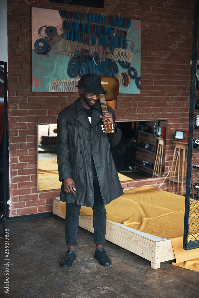 Musician in black trench coat and bucket standing with guitar. Songwriter in loft room. Fashion 2019 Musician in black trench coat and bucket standing with guitar. Songwriter in loft room. Fashion 2019
