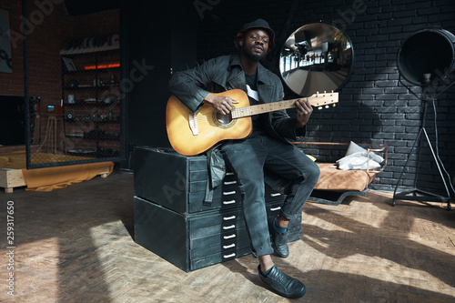 Musician in black trench coat and bucket hat sitting on bed and playing guitar. Songwriter in loft room. Fashion 2019 photo