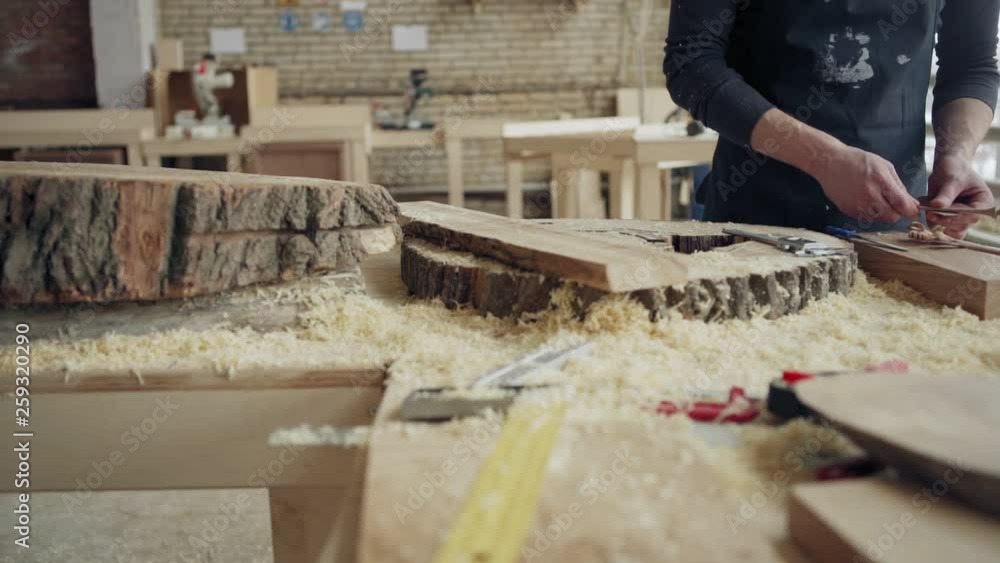 Hands of unrecognizable craftsman using wood file to make decorated hair sticks, his workbench covered with wood slices, tools and sawdust; tracking slow motion shot Hands of unrecognizable craftsman using wood file to make decorated hair sticks, his workbench covered with wood slices, tools and sawdust; tracking slow motion shot