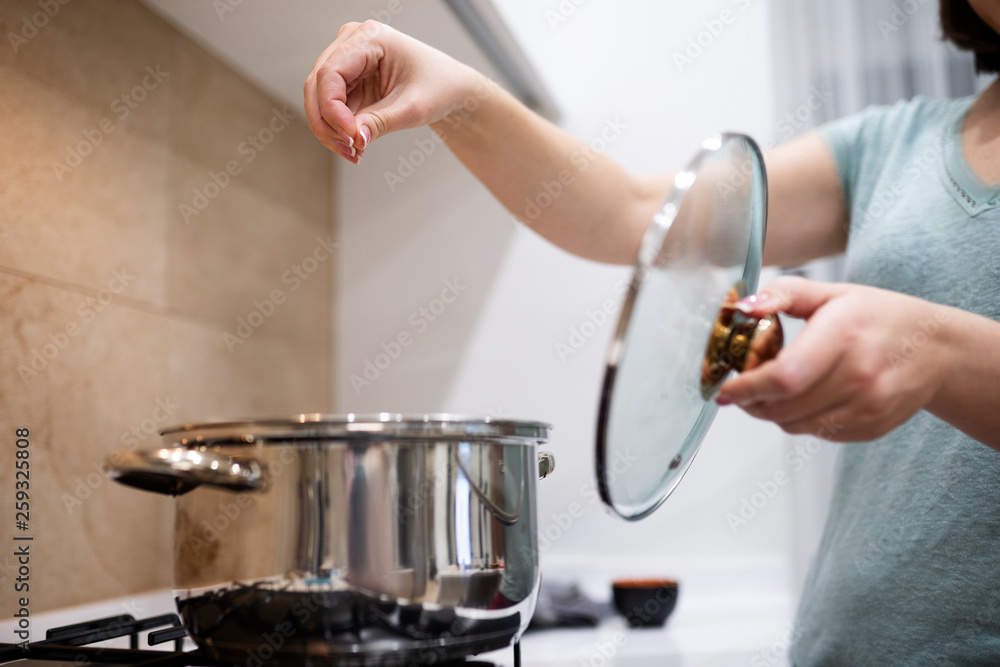 Beautiful young woman housewife prepairing dinner, pouring salt in big steel saucepan, standing on gas-stove.