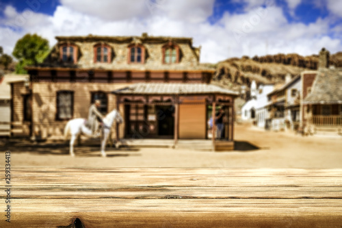 Wooden old table of free space for your decoration and wild west blurred background of big building and cowboy on horse.  © magdal3na