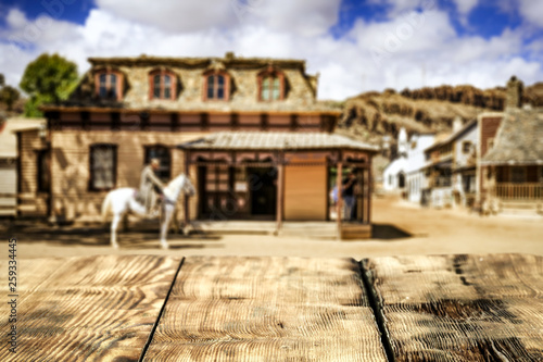 Wooden old table of free space for your decoration and wild west blurred background of big building and cowboy on horse.  © magdal3na