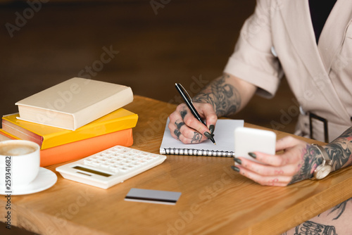 partial view of tattooed businesswoman writing in notebook and holding smartphone