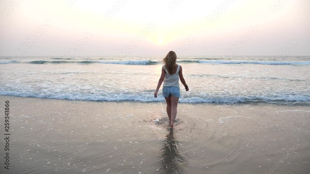 Young slender girl barefoot go into the sea at sunset and spray flying from the waves. Happy woman resting on the sea on vacation. Young slender girl barefoot go into the sea at sunset and spray flying from the waves. Happy woman resting on the sea on vacation.