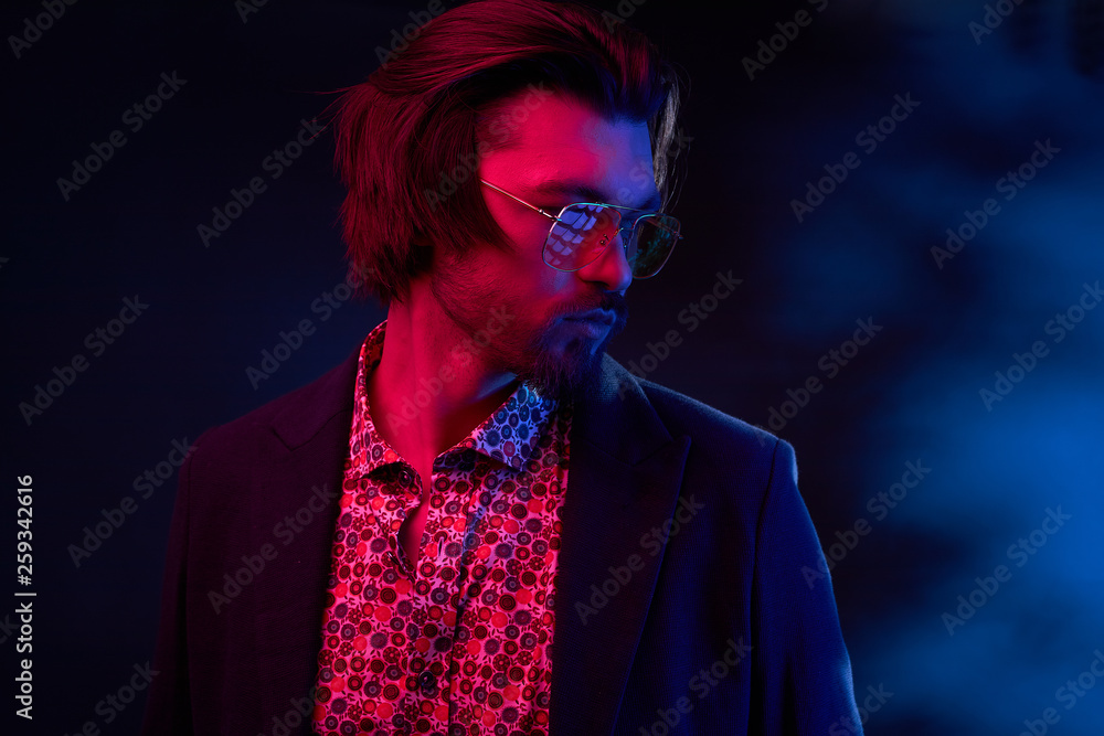 Neon light portrait of handsome male model with dark medium length hair, mustaches and beard in sunglasses, weared in dark jacket and floral shirt. Neon light portrait of handsome male model with dark medium length hair, mustaches and beard in sunglasses, weared in dark jacket and floral shirt.