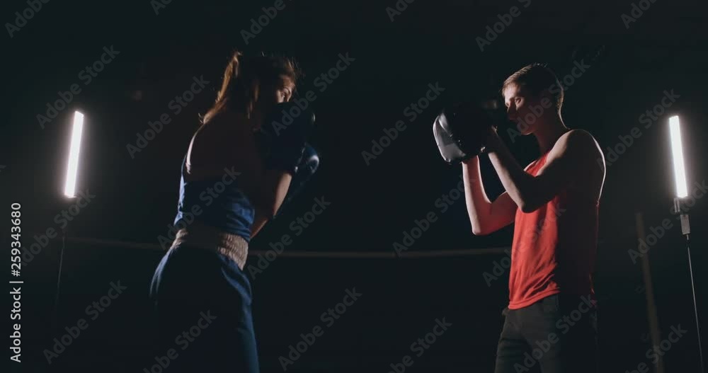 Beautiful brunette boxer working out blows to the paws with a trainer in a dark room