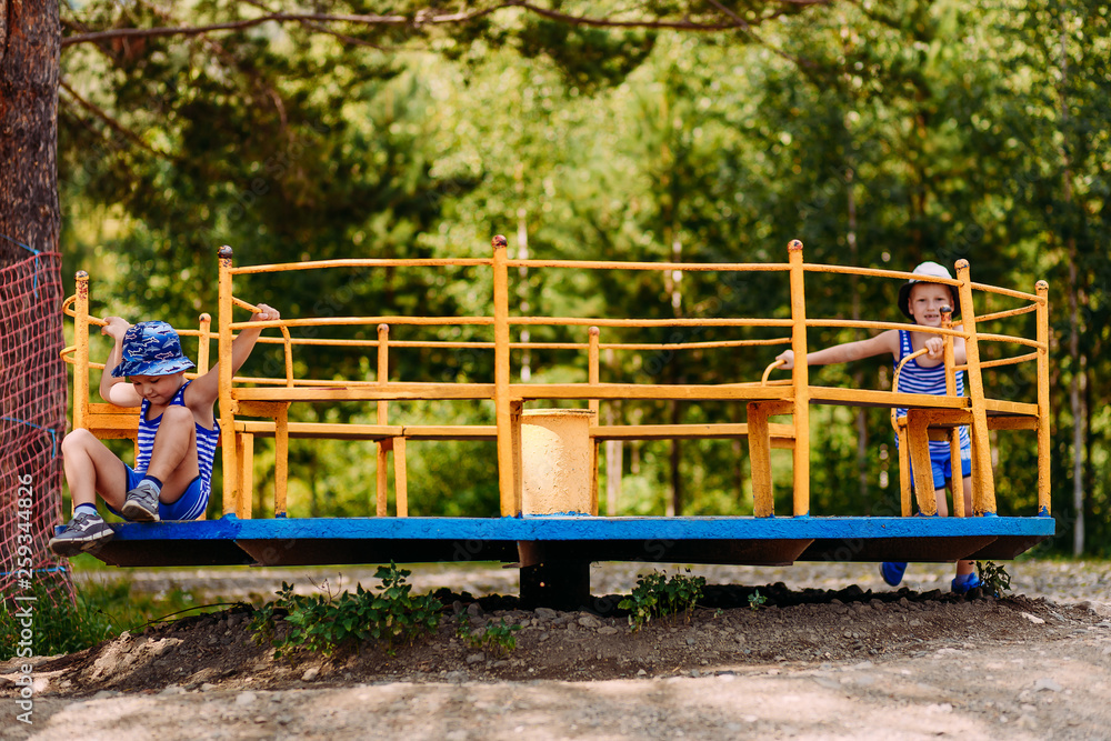 two cheerful boys in hats and t-shirts play on a big iron carousel on the Playground two cheerful boys in hats and t-shirts play on a big iron carousel on the Playground