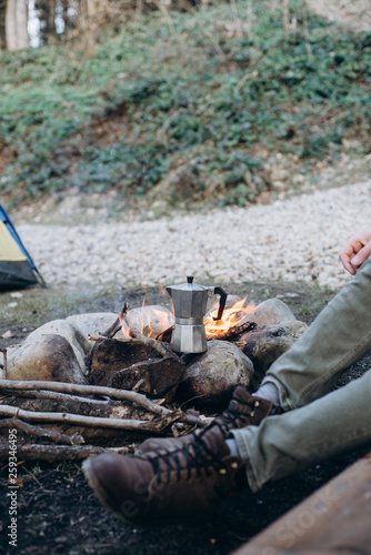 Cropped outdoor image of young man explorer sitting near to bonfire preparing hot beverage in mountains. Concept adventure active vacations. Summer camping
