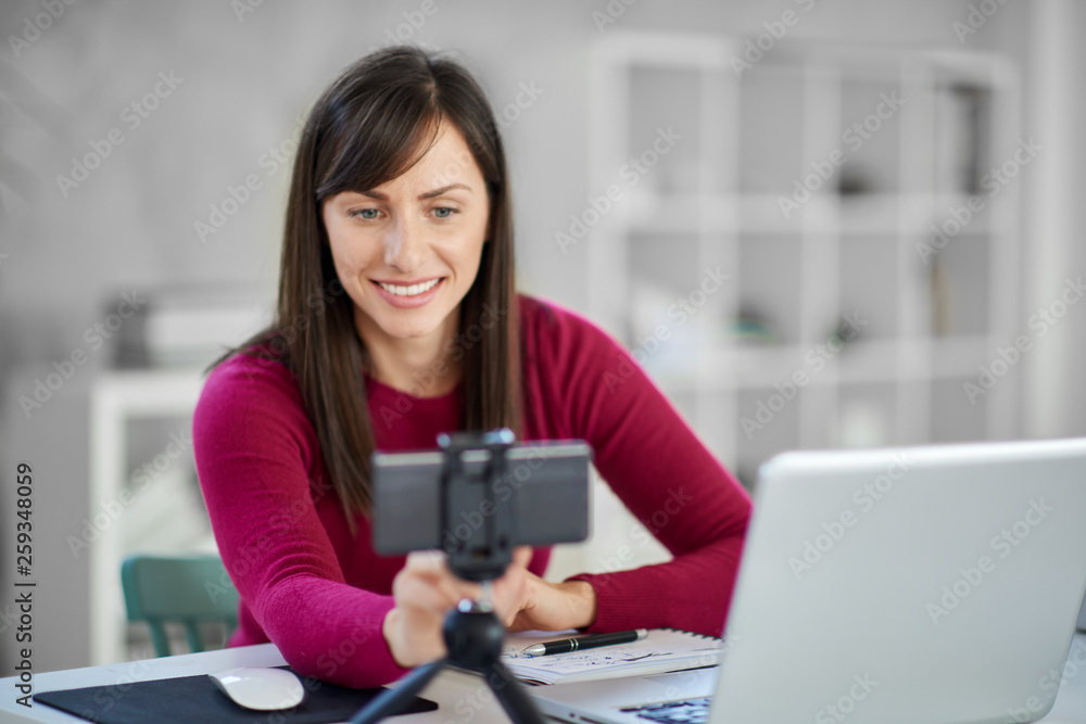 Charming Caucasian brunette dressed casual adjusting smart phone for video call. In front of her laptop, office interior. Charming Caucasian brunette dressed casual adjusting smart phone for video call. In front of her laptop, office interior.