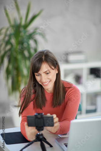 Charming Caucasian brunette dressed casual adjusting smart phone for video call. In front of her laptop, office interior.