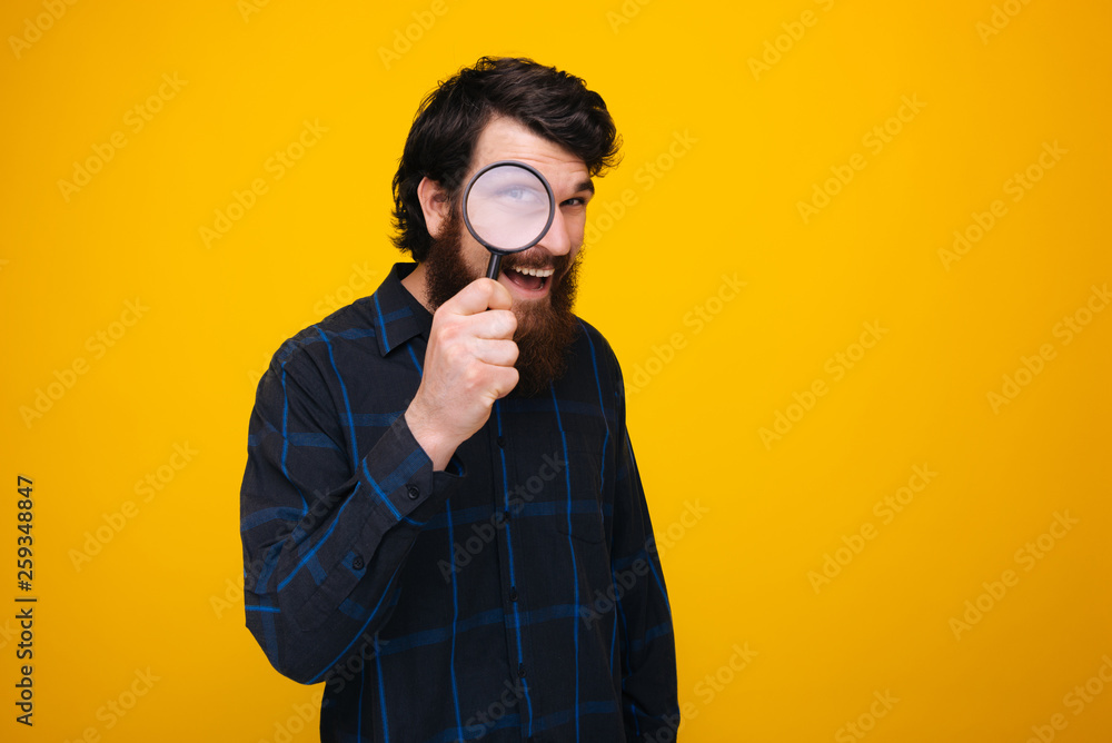 Young bearded man smiling and looking with an eye through a magnifying glass on yellow background. Young bearded man smiling and looking with an eye through a magnifying glass on yellow background.