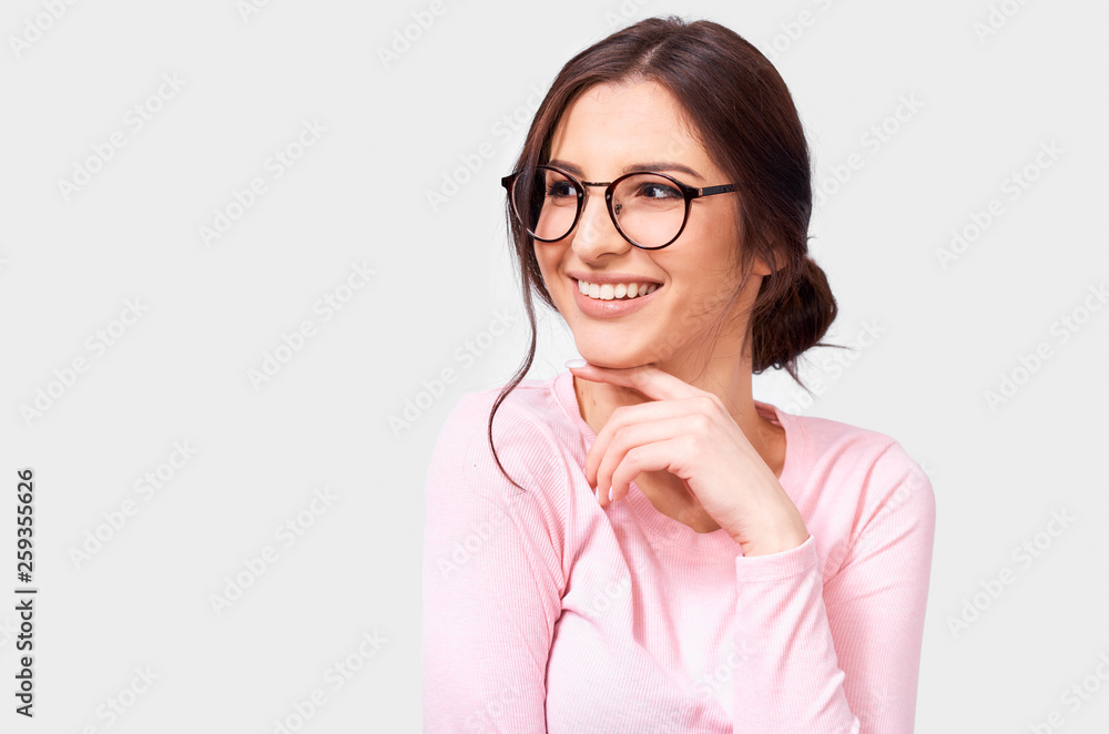 Portrait of pretty positive European young woman in pink long sleeve blouse and round transparent eyewear, smiling broadly and looking aside, isolated over white background. People real emotions Portrait of pretty positive European young woman in pink long sleeve blouse and round transparent eyewear, smiling broadly and looking aside, isolated over white background. People real emotions