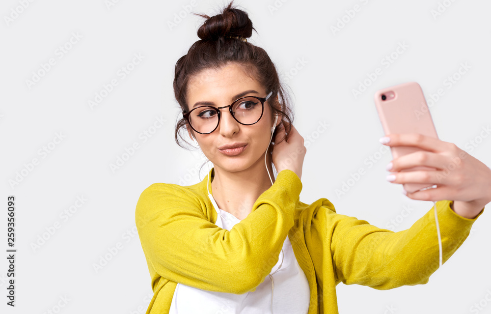 Shot of young European woman student in casual clothes, taking self portrait over white studio wall. Happy female posing and taking a selfie on her smart phone, isolated over gray background.
