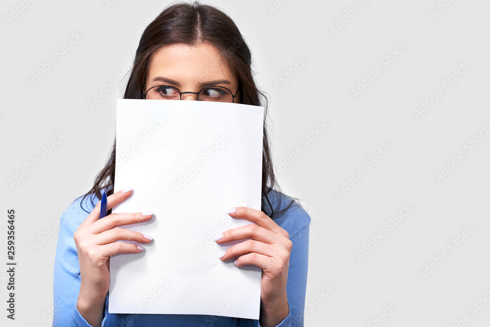 Portrait of pretty brunette young woman student wearing blue blouse and round transparent eyeglasses, hiding with white paper on her face, looking aside. Cheerful female office worker on white wall. Portrait of pretty brunette young woman student wearing blue blouse and round transparent eyeglasses, hiding with white paper on her face, looking aside. Cheerful female office worker on white wall.